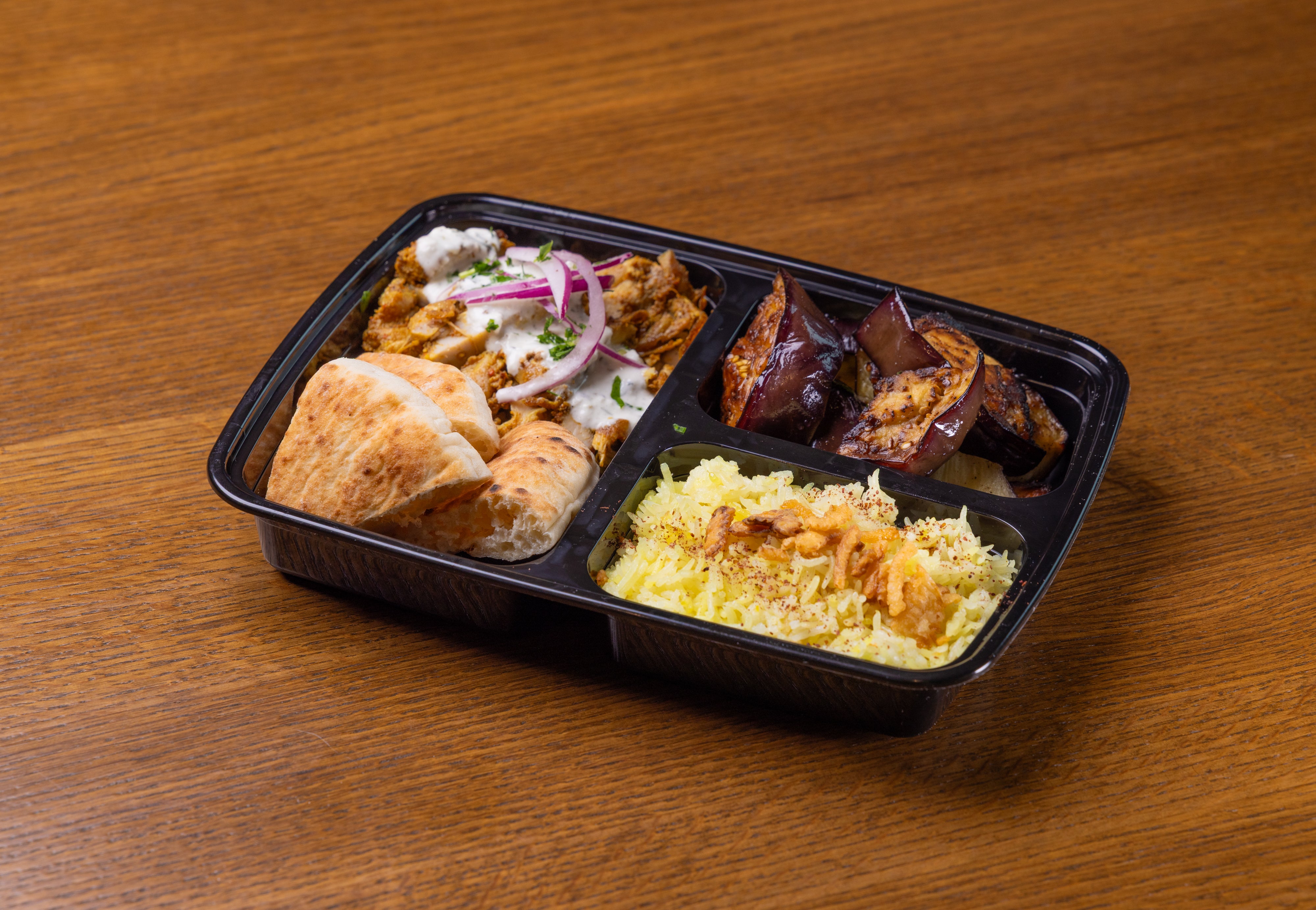 A black to-go box with chicken shawarma and tzatziki sauce, pita wedges, roasted eggplant and saffron basmati rice.