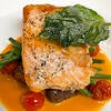 Pan-seared salmon fillet topped with a crispy fried basil leaf, presented on a white plate. It is served over a bed of green beans and roasted cherry tomatoes, all resting on a vibrant orange sauce.