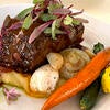 Slow-braised beef short rib topped with a rich red wine demi-glaze and garnished with fresh greens, served with roasted baby carrots, creamy mashed potatoes and glazed baby onions on a white plate.