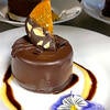 A chocolate mousse cake glazed in chocolate and topped with a candied orange, gold leaf, and sea salt on a white plate.